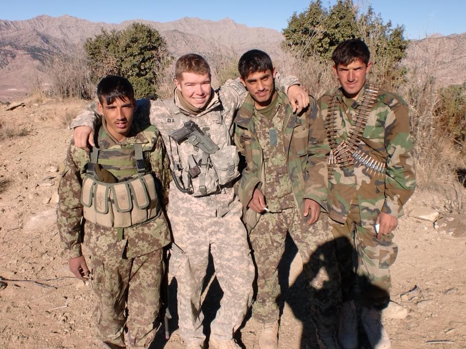 Ken Clark, arms around three Afghan National Security Forces soldiers on a hillside above a valley, all four of them grinning.