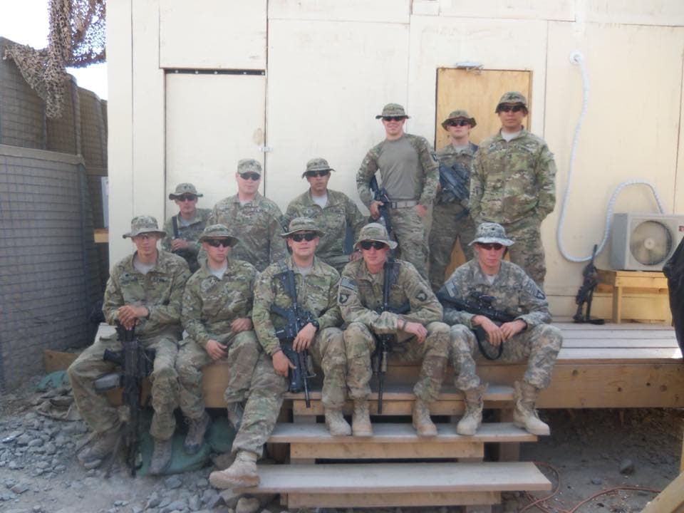Eleven soldiers in combat uniform grouped on the steps of a plywood building at a combat outpost, HESCO barriers and camouflage netting behind them.
