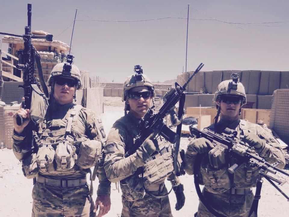 Three soldiers in full body armor and helmets standing inside an outpost, weapons up, HESCO walls behind them.