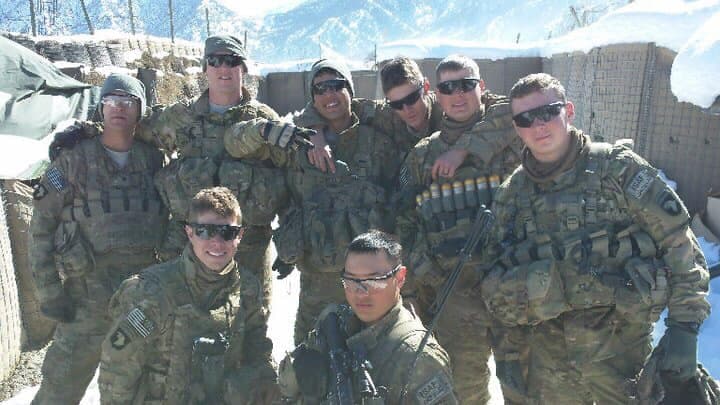 Eight soldiers in winter combat gear grouped at a snow-covered outpost, mountains rising behind the perimeter wall.